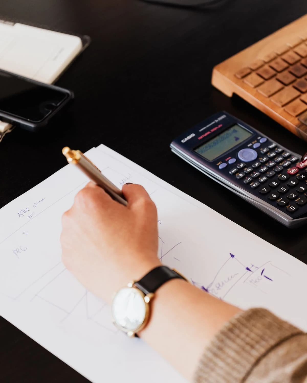 Mână care notează calcule pe o foaie, lângă un calculator de birou, fotografie ilustrativă.